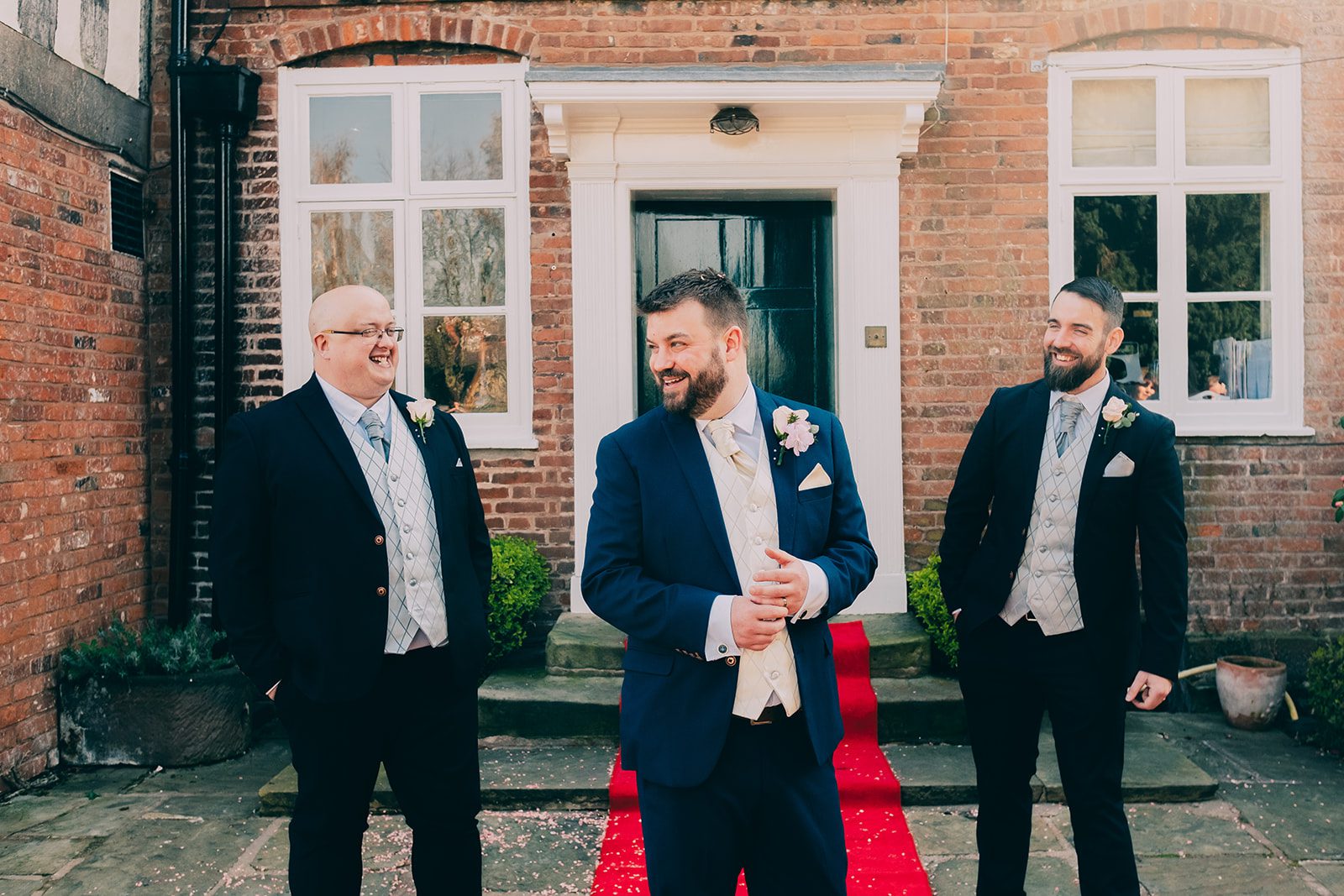Groom and groomsmen in suits, laughing at each other at The Moat House in Acton Trussell